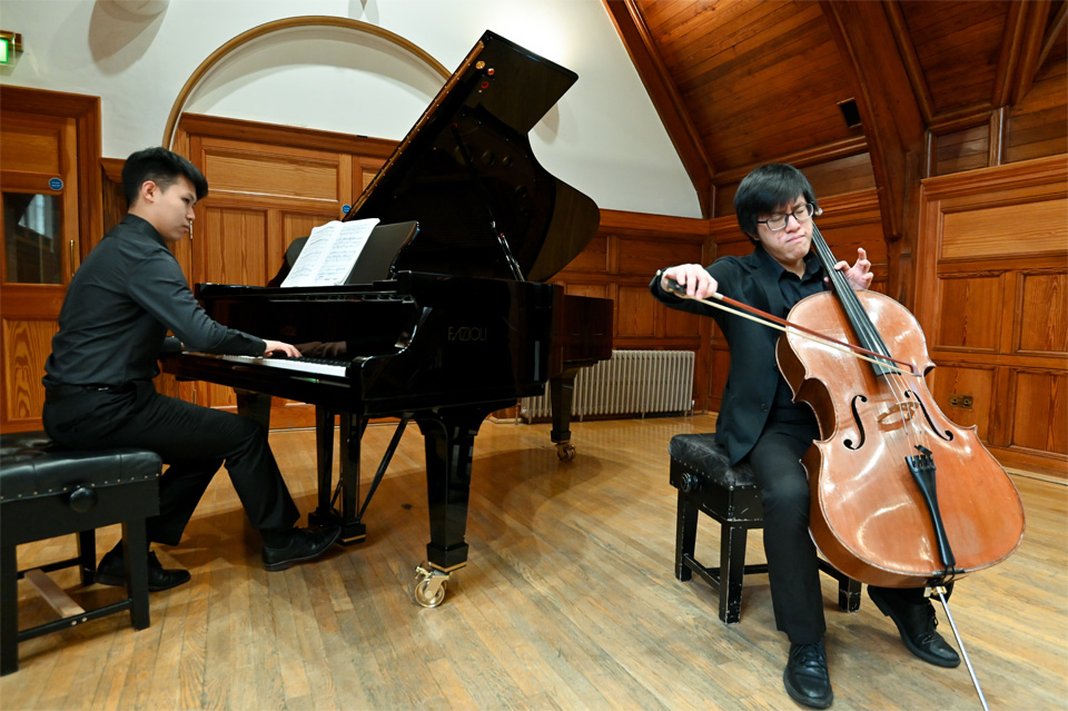 An Asian male student, playing the cello, with another student accompanying him on the piano, in a wood-panelled room.